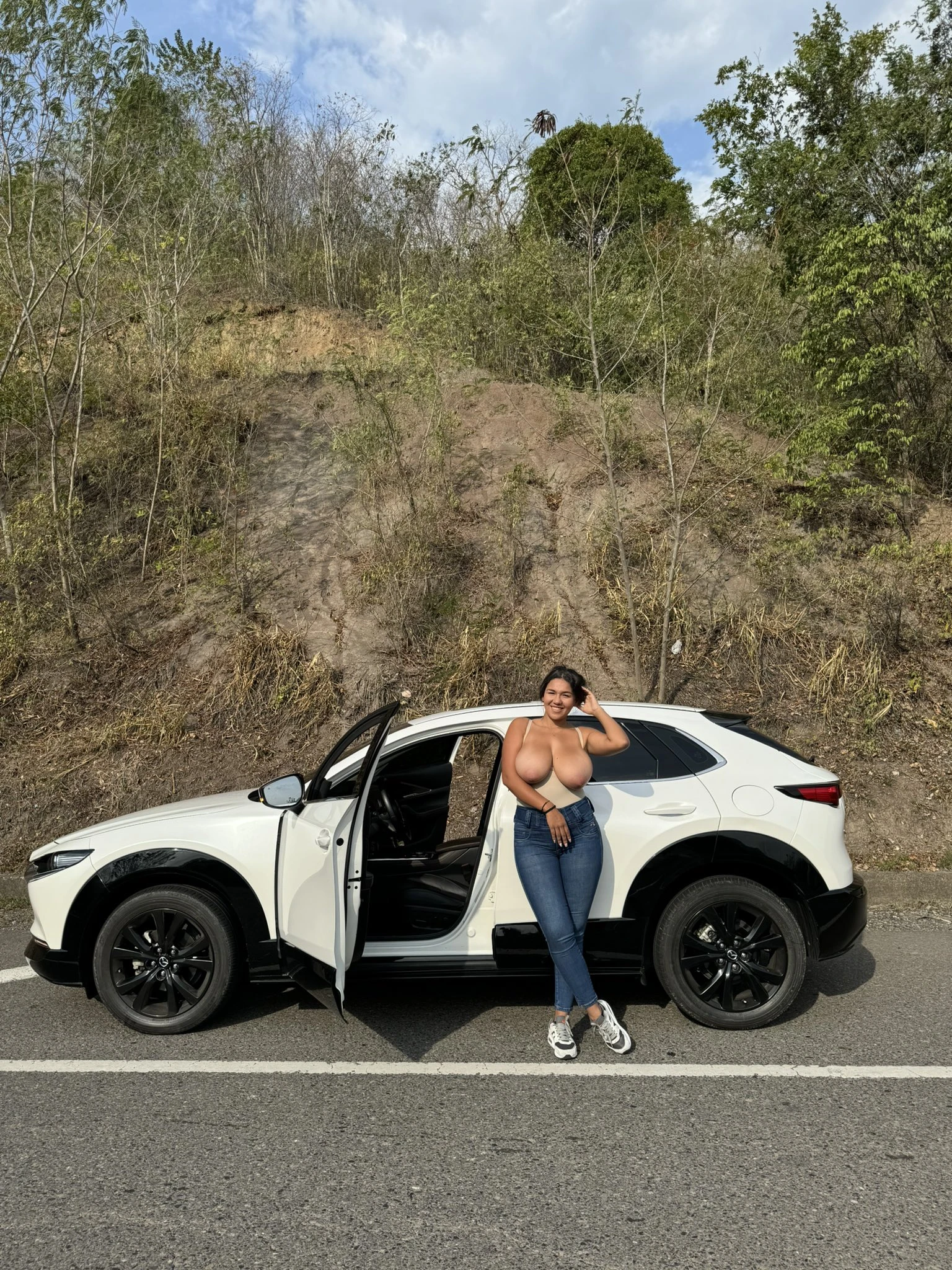 Uninhibited MILF beauty in denim pants shows her topless big tits while standing near a car in the middle of the road