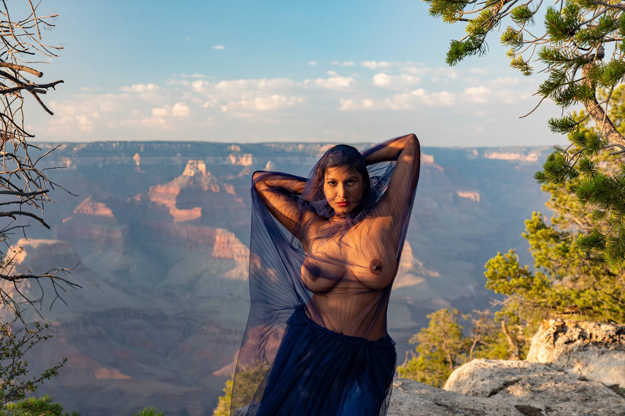 Desi model showing her big breasts wearing a sheer, dark blue scarf, standing on a cliff with hands behind her head, with Grand Canyon in background