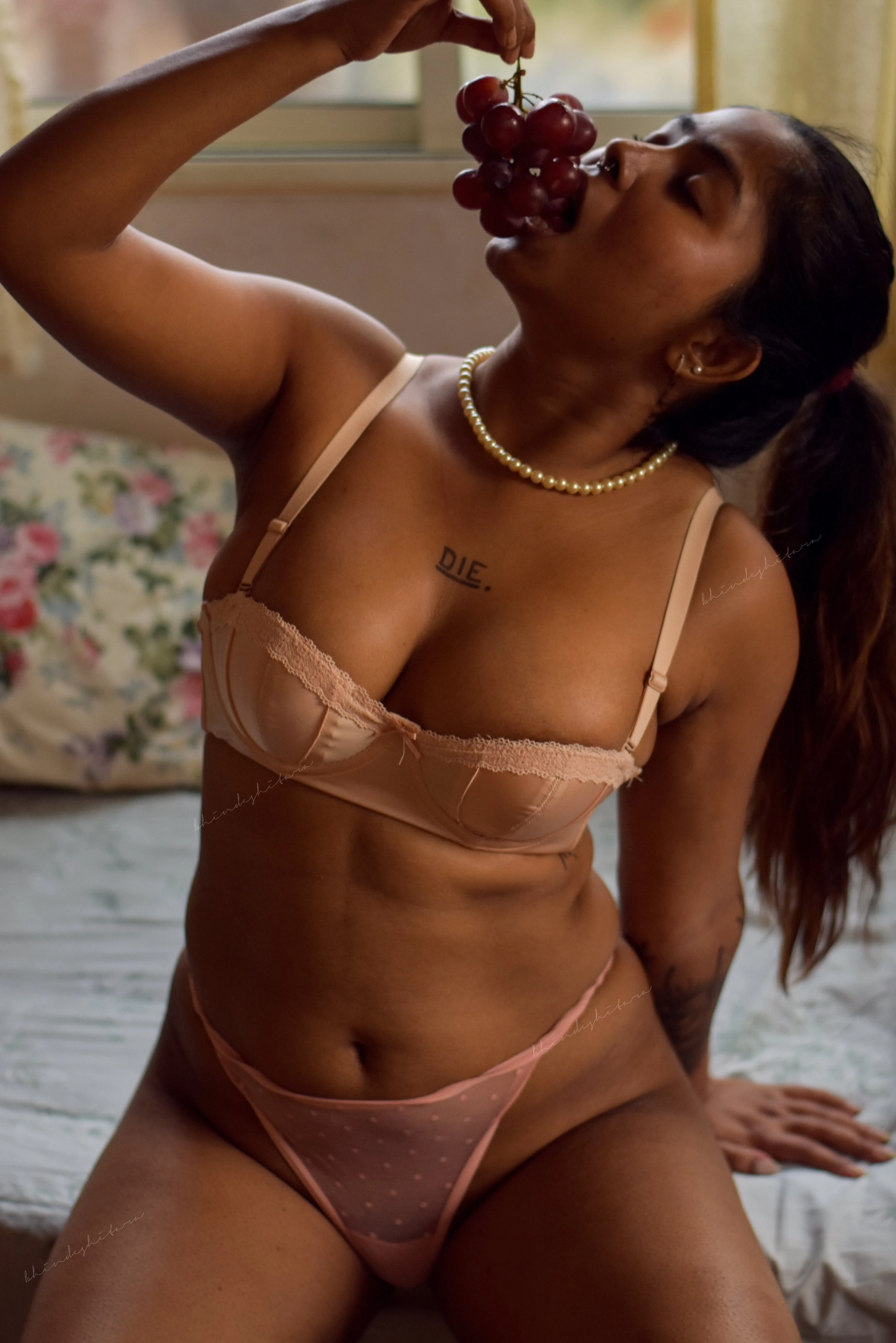 Desi MILF sitting on a bed wearing a pink lace bra and panties holding a bunch of grapes