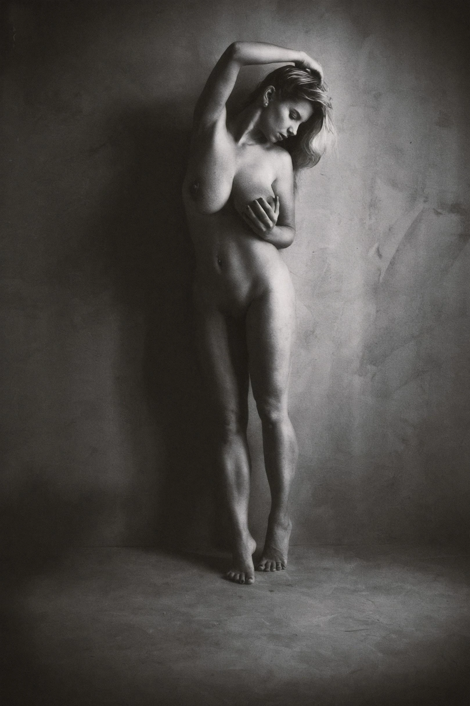 Katerina Hartlova, stands nude gracefully against a textured wall, her pose conveying both vulnerability and strength in a black and white scene