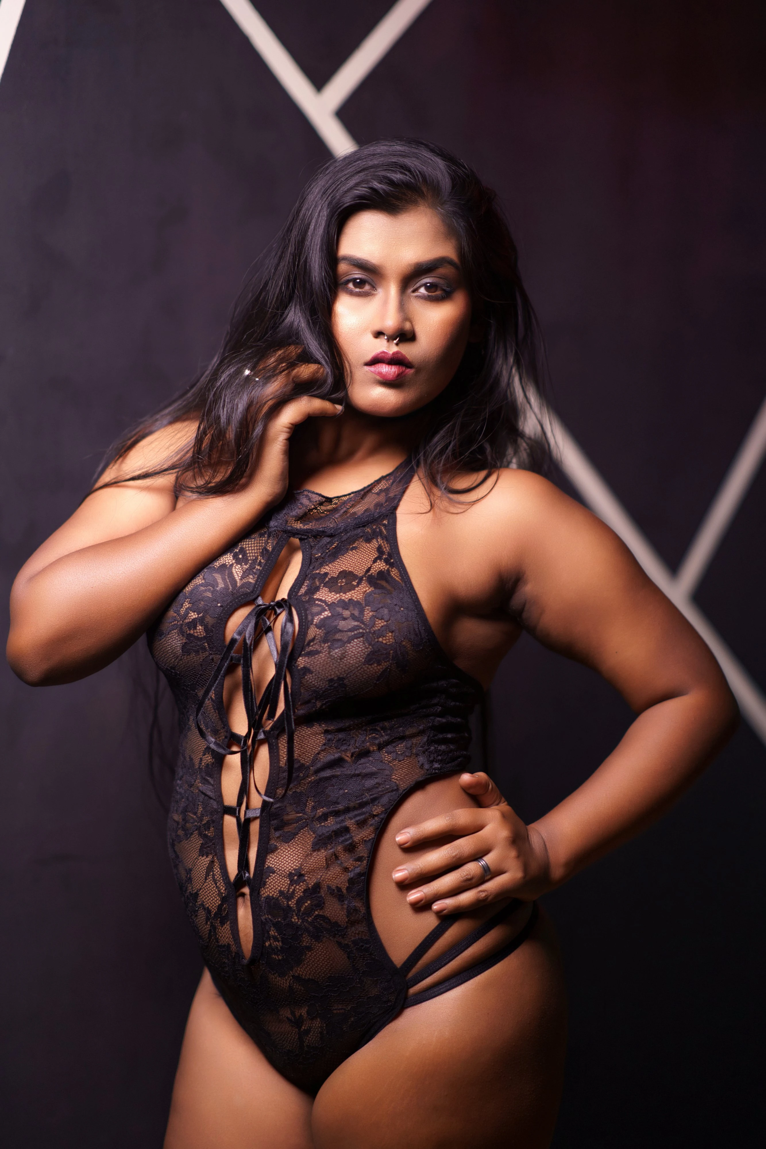 Curvy Desi babe in a black lace bodysuit with floral patterns, showing off her large tits and confidently staring into the camera