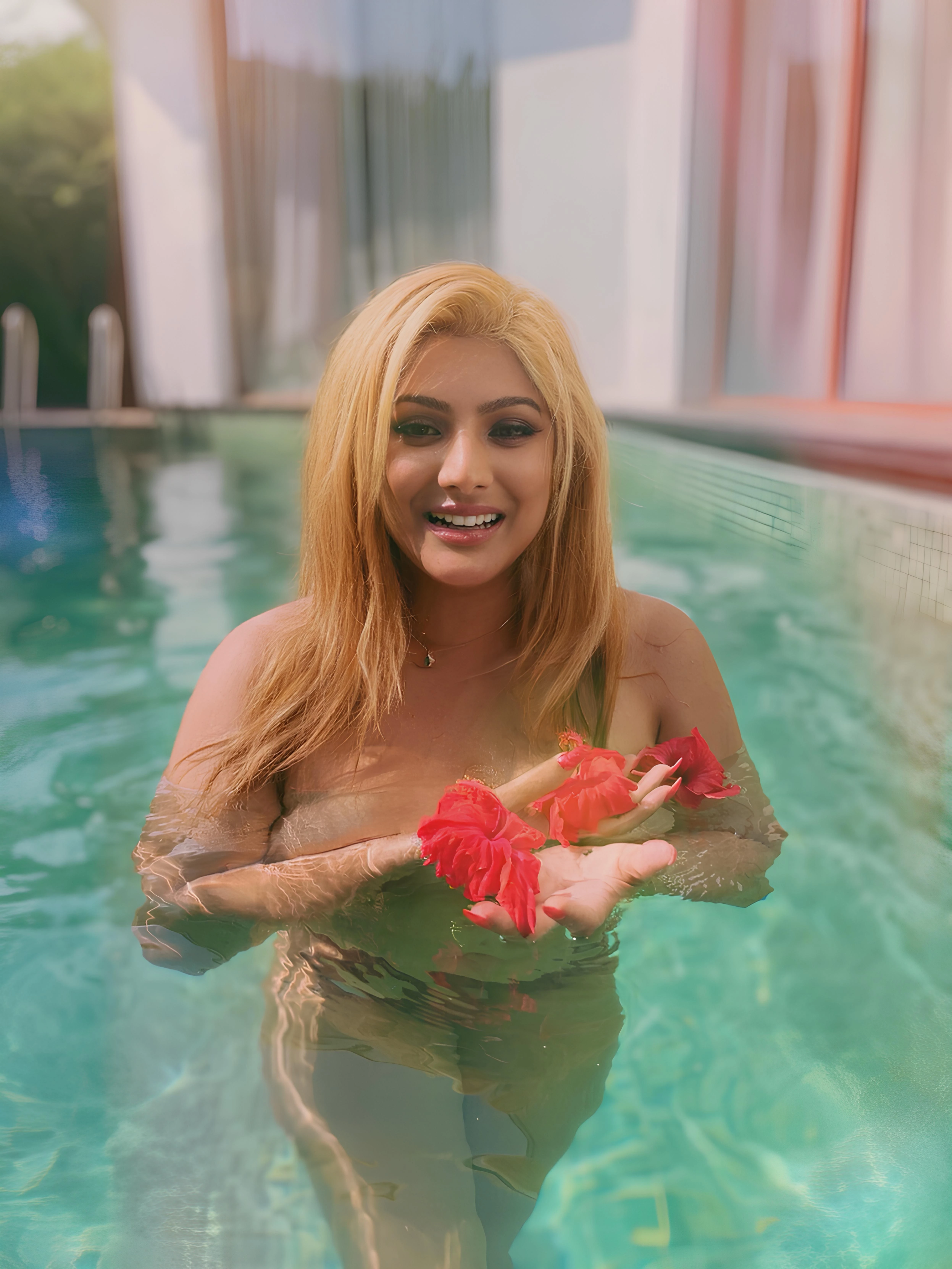 Sultry Desi actress stands waist-deep in a pool, holding red flower petals, exuding a cheerful and summery vibe