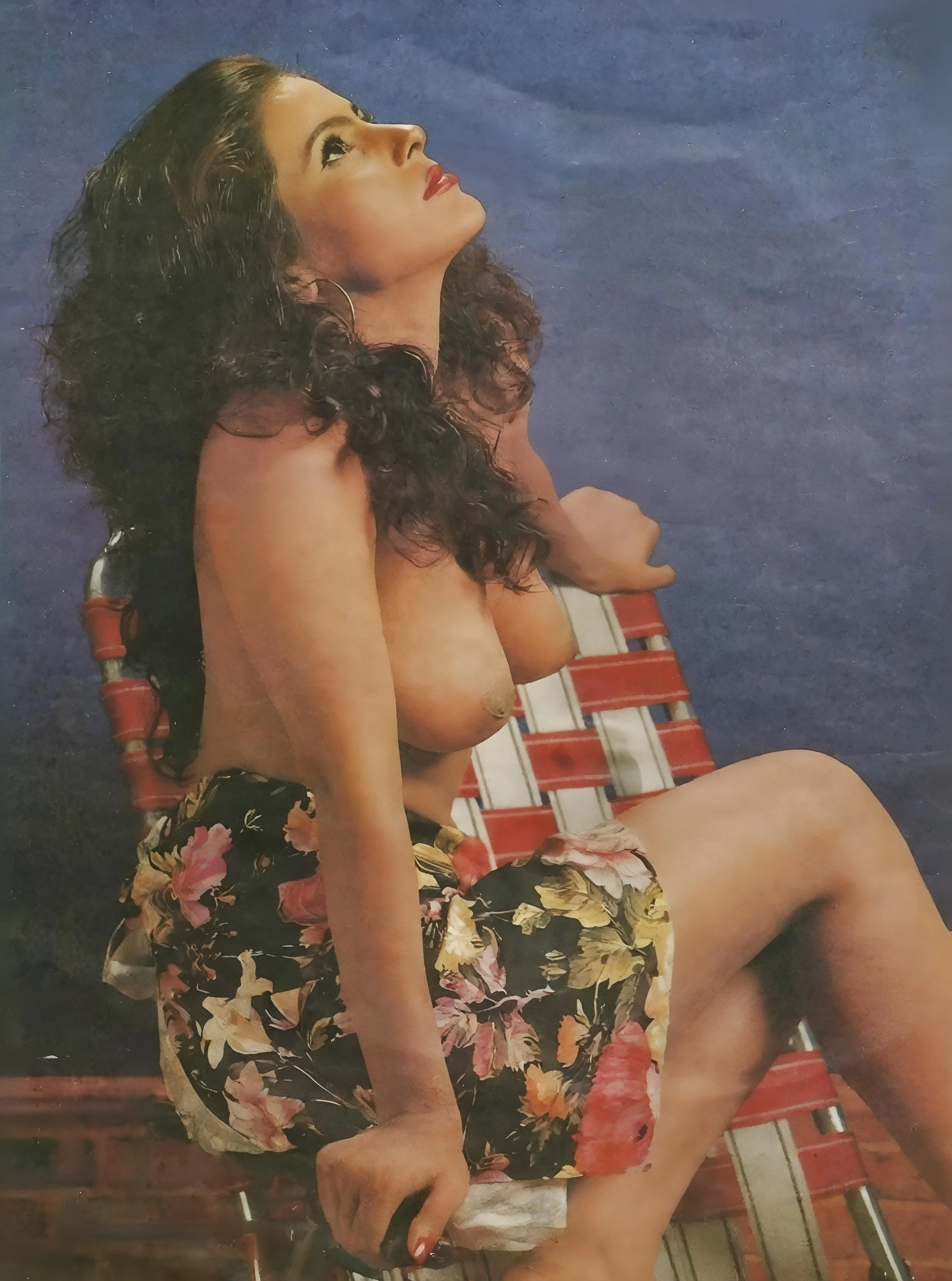 Desi model from a vintage porn magazine sits confidently in a woven chair, her topless form and upward gaze creating a timeless, alluring scene