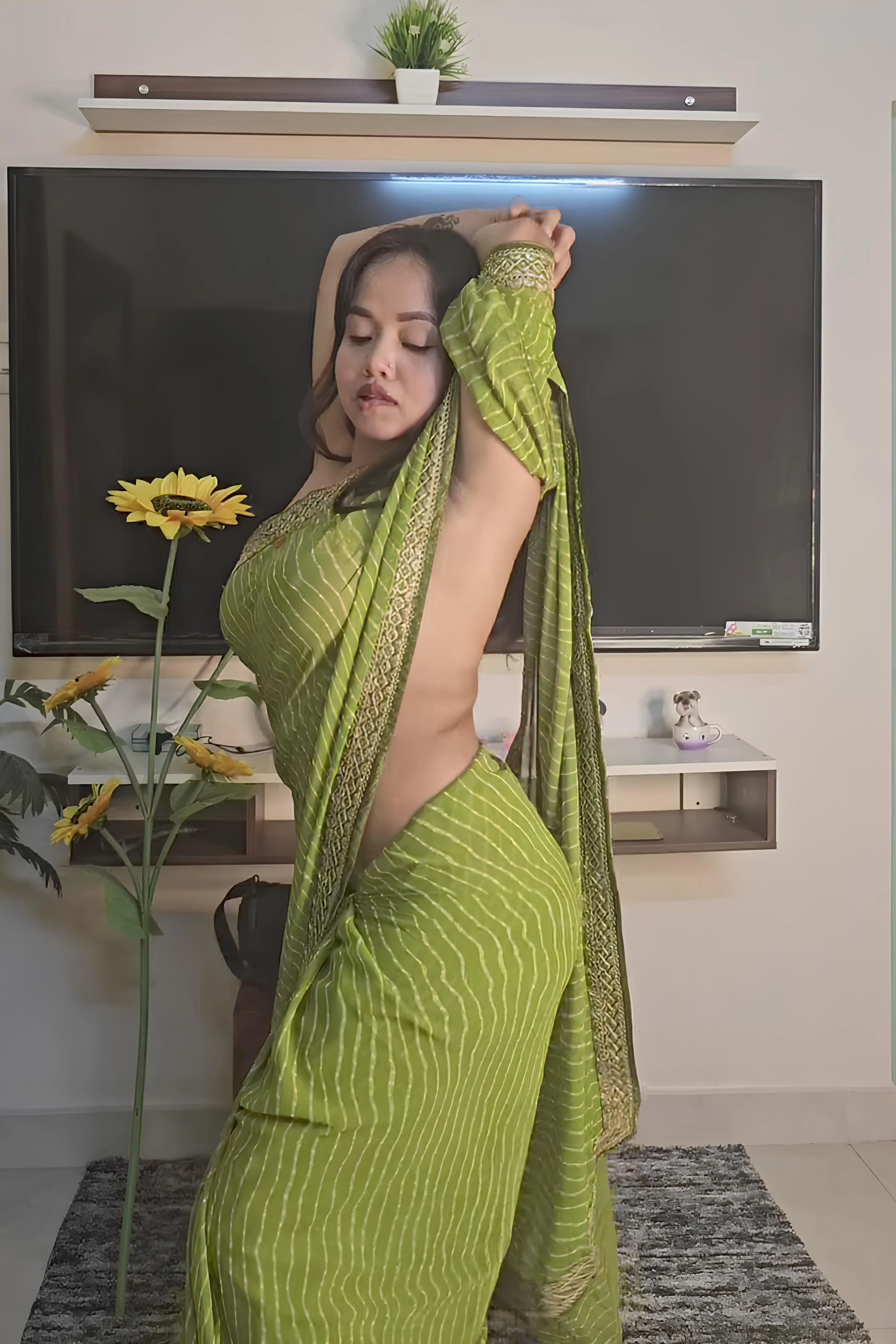 Desi DK Insane in a vibrant green braless saree with white stripes, revealing her bare back and side in a modern living room