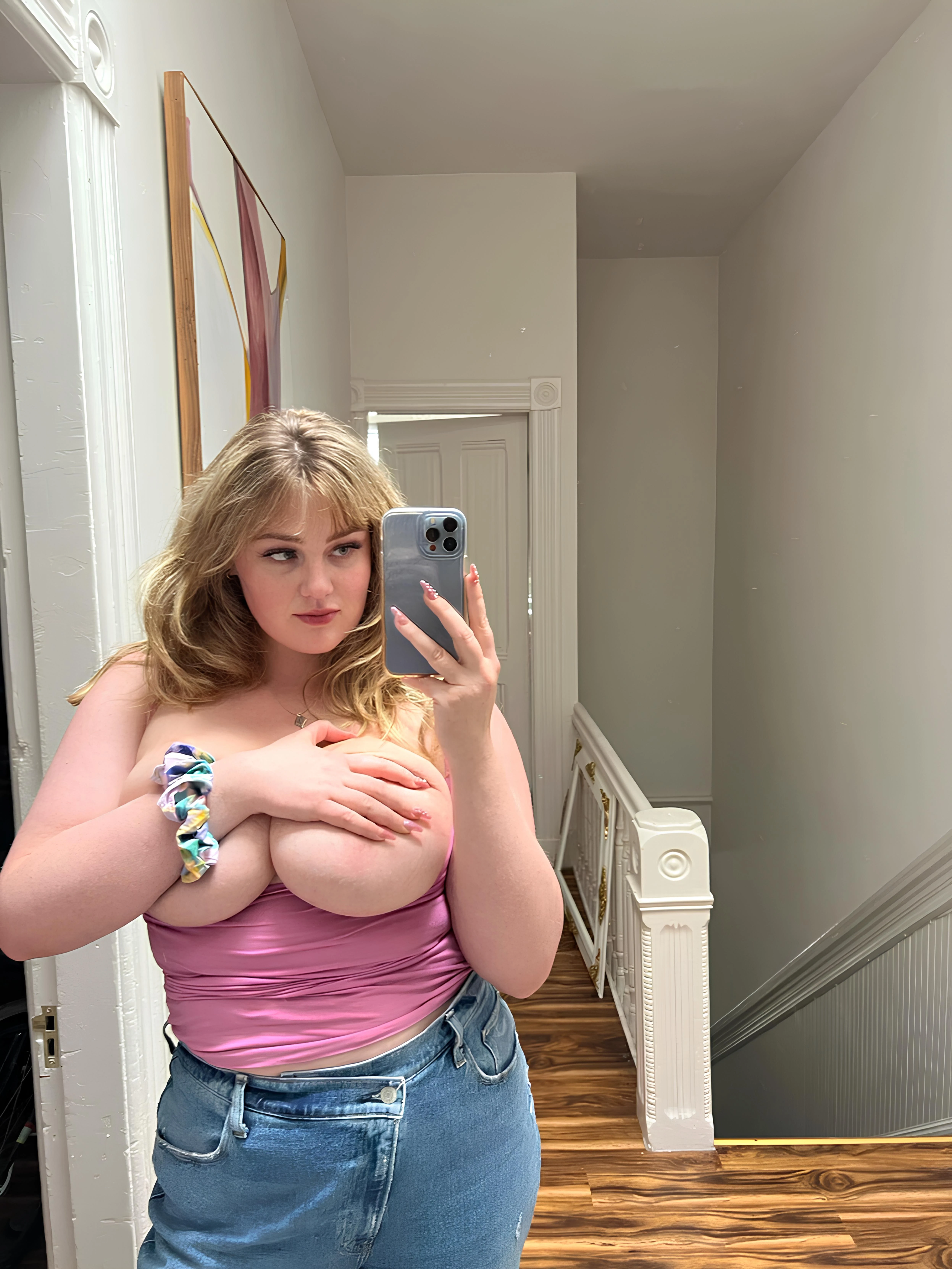 Blonde vixen in a tight pink top takes a selfie, her large, round breasts and pink nipples hidden by her hand in a narrow hallway