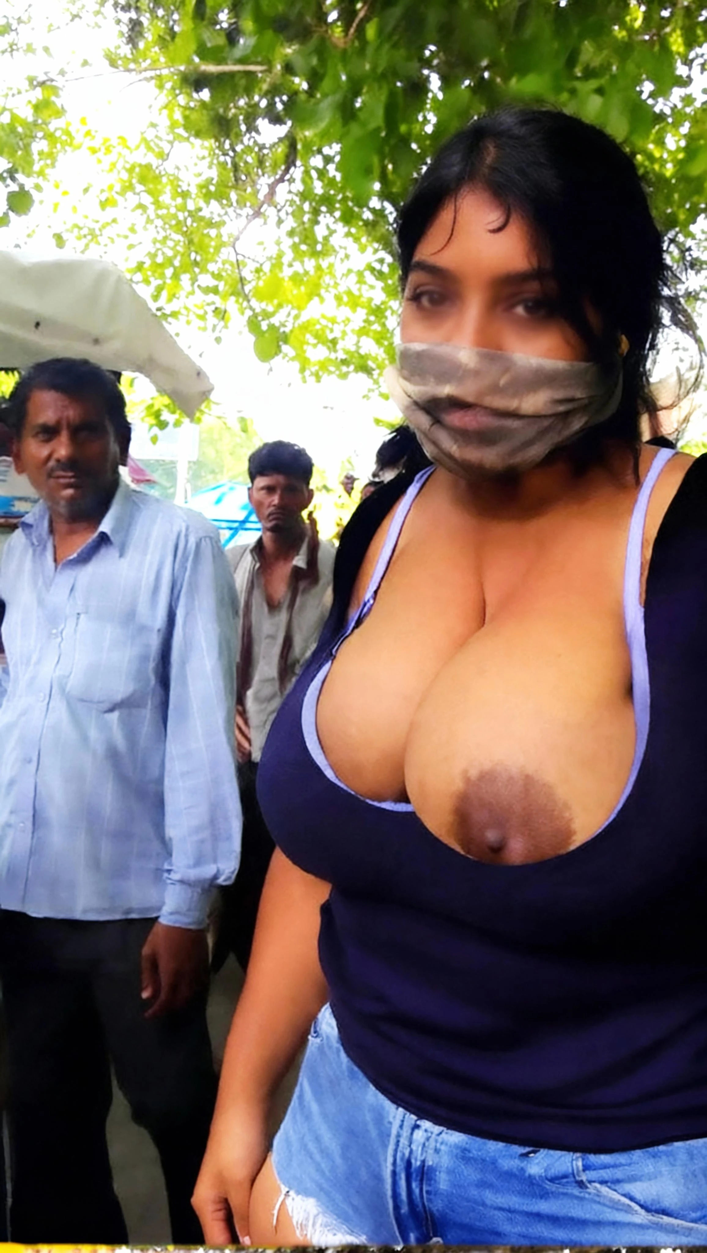 Exhibitionist Desi woman pulling down dark tank top exposing massive DD tits with large dark brown areolas in public with men background