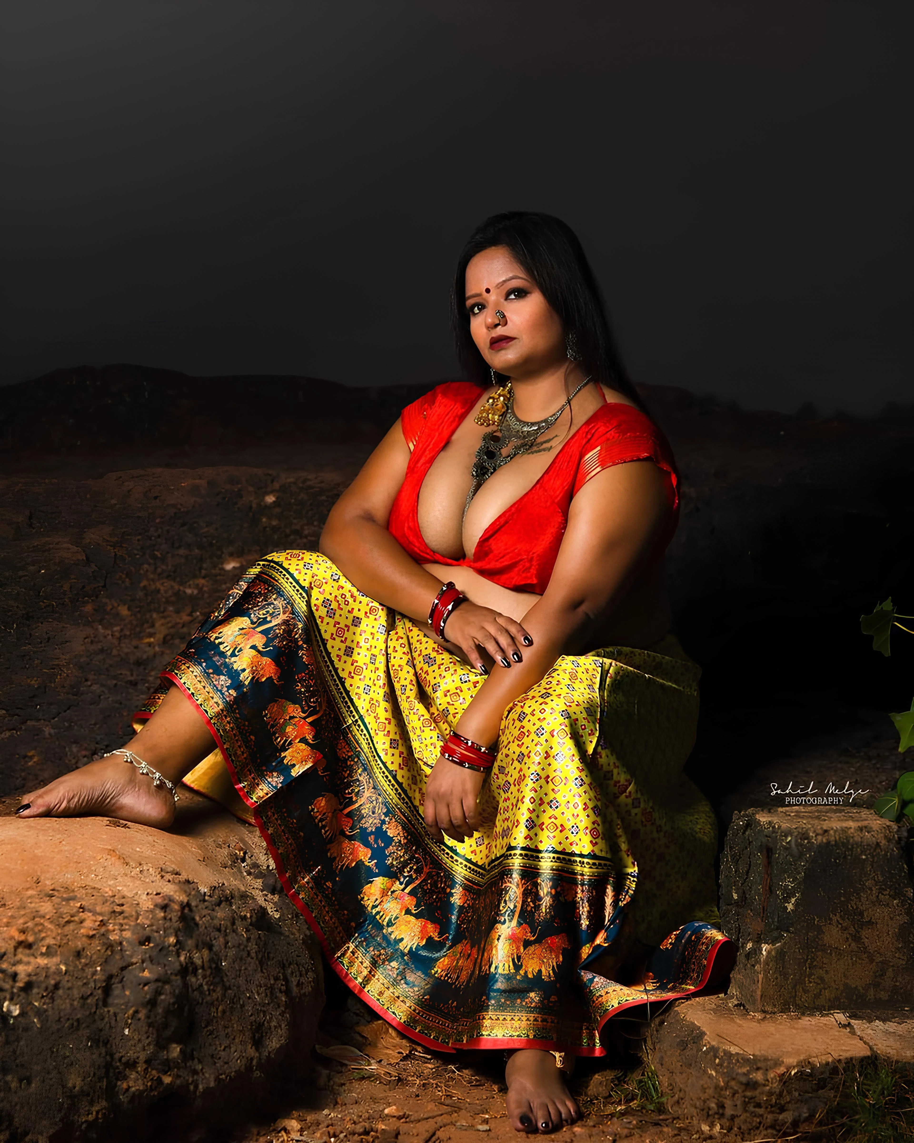 Desi MILF seated on rocky terrain, her substantial bare D-cup breasts with large dark pink areolas spilling from a plunging red blouse under a dusky sky