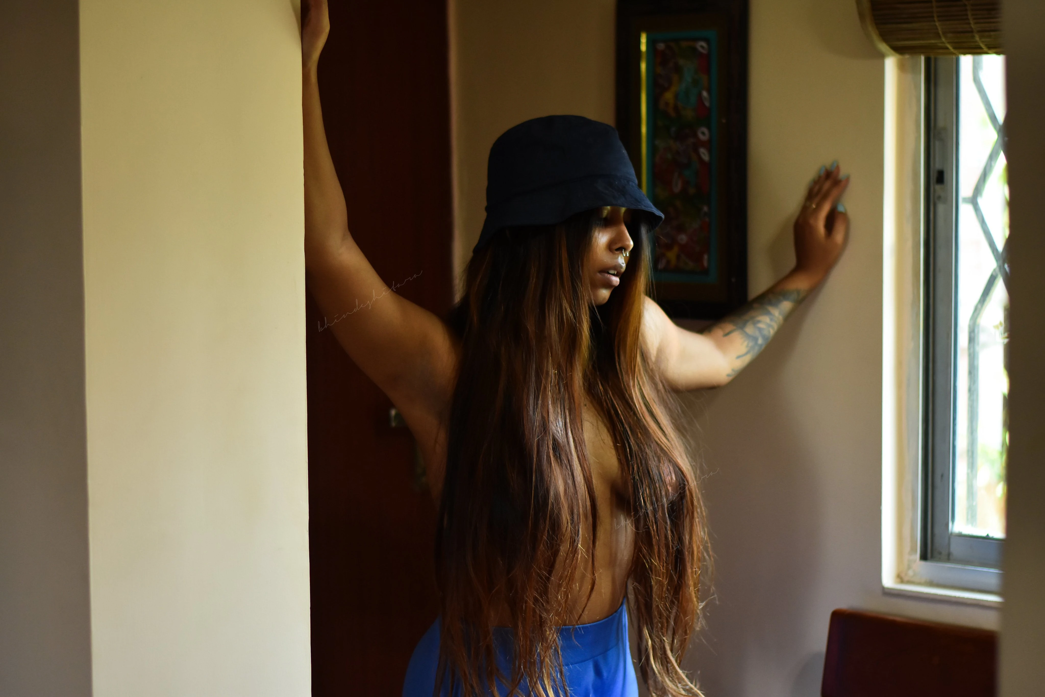 Bhindeshitara topless indoors wearing a dark blue bucket hat, her bare torso visible as she leans against a pillar with arms raised