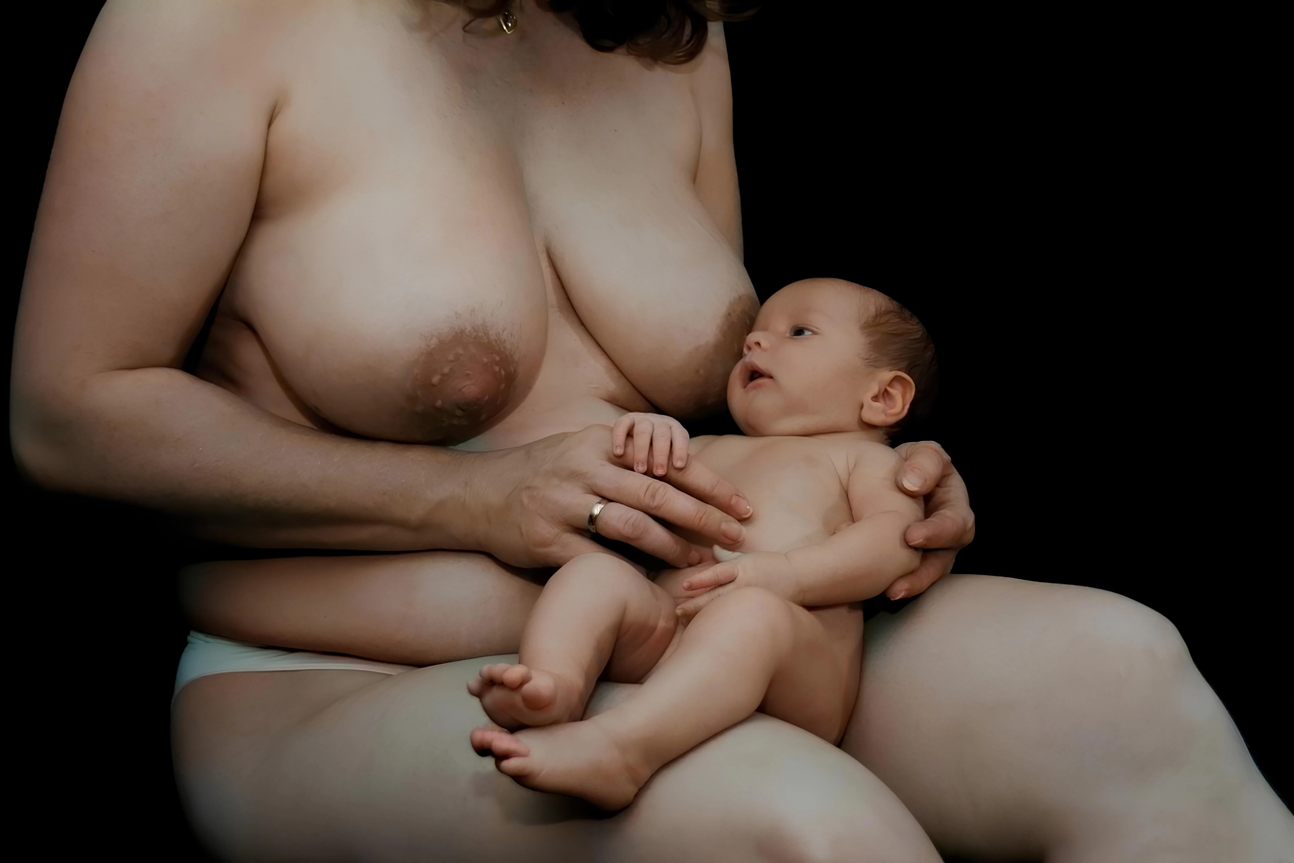 Topless woman with large full breasts and dark areolas sitting indoors, holding a baby against her chest in soft lighting