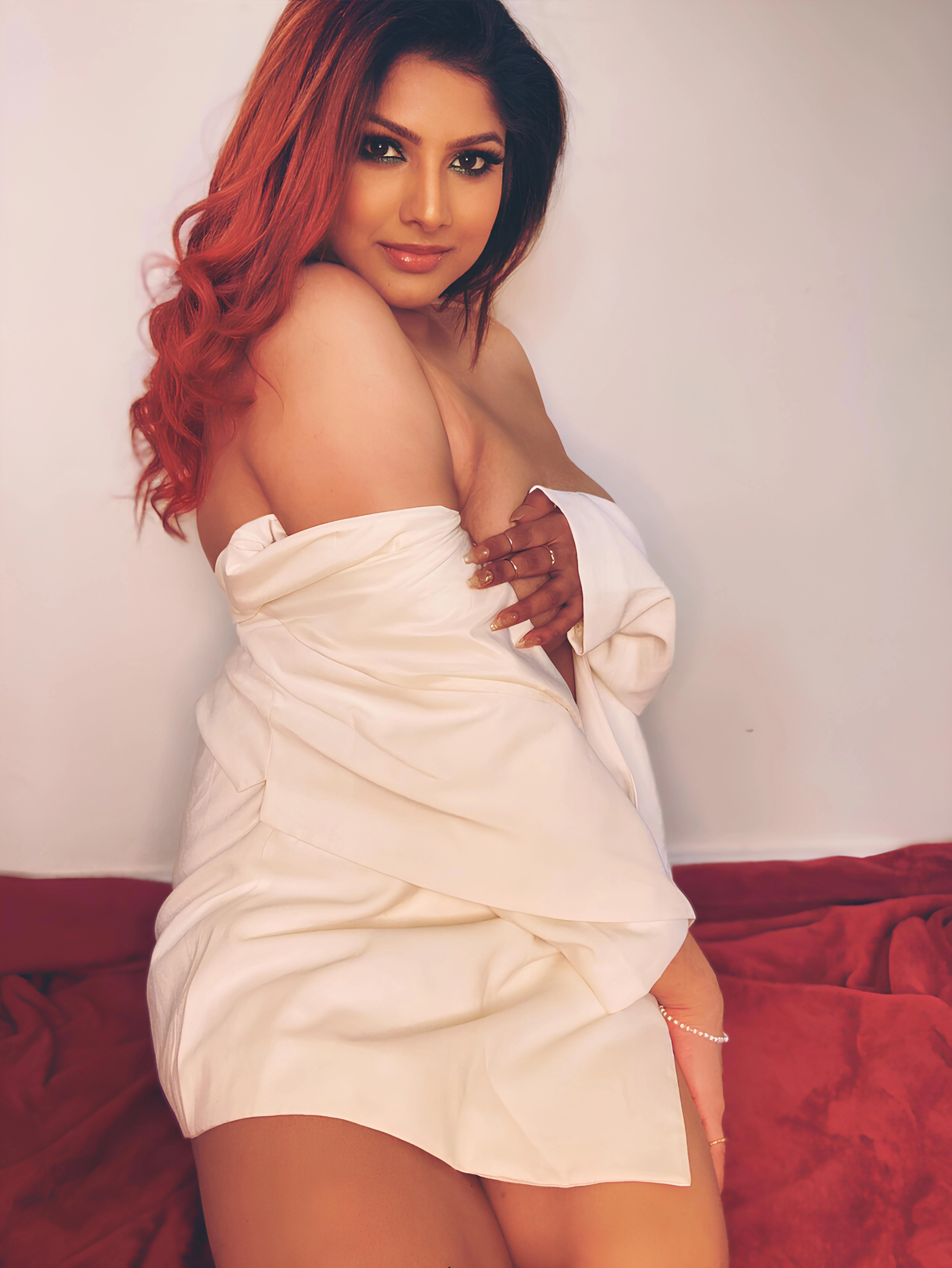 Joshna Fernando seated on red fabric wearing a white draped garment, her full left breast exposed while her hand partially covers the right