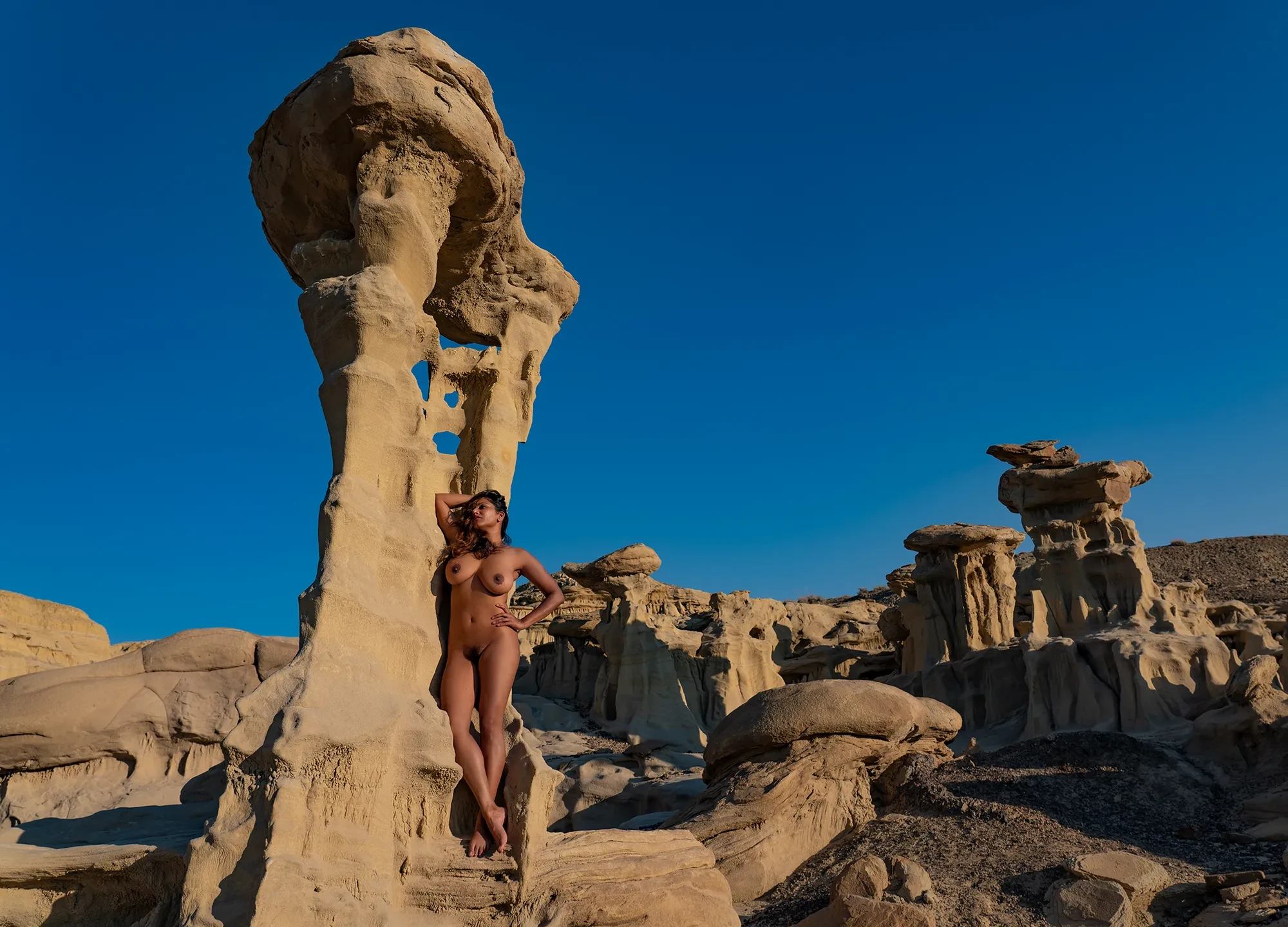 Dakini completely naked in a desert landscape with rock formations, her B-cup breasts with pale pink nipples and dark pubic hair visible under bright sky