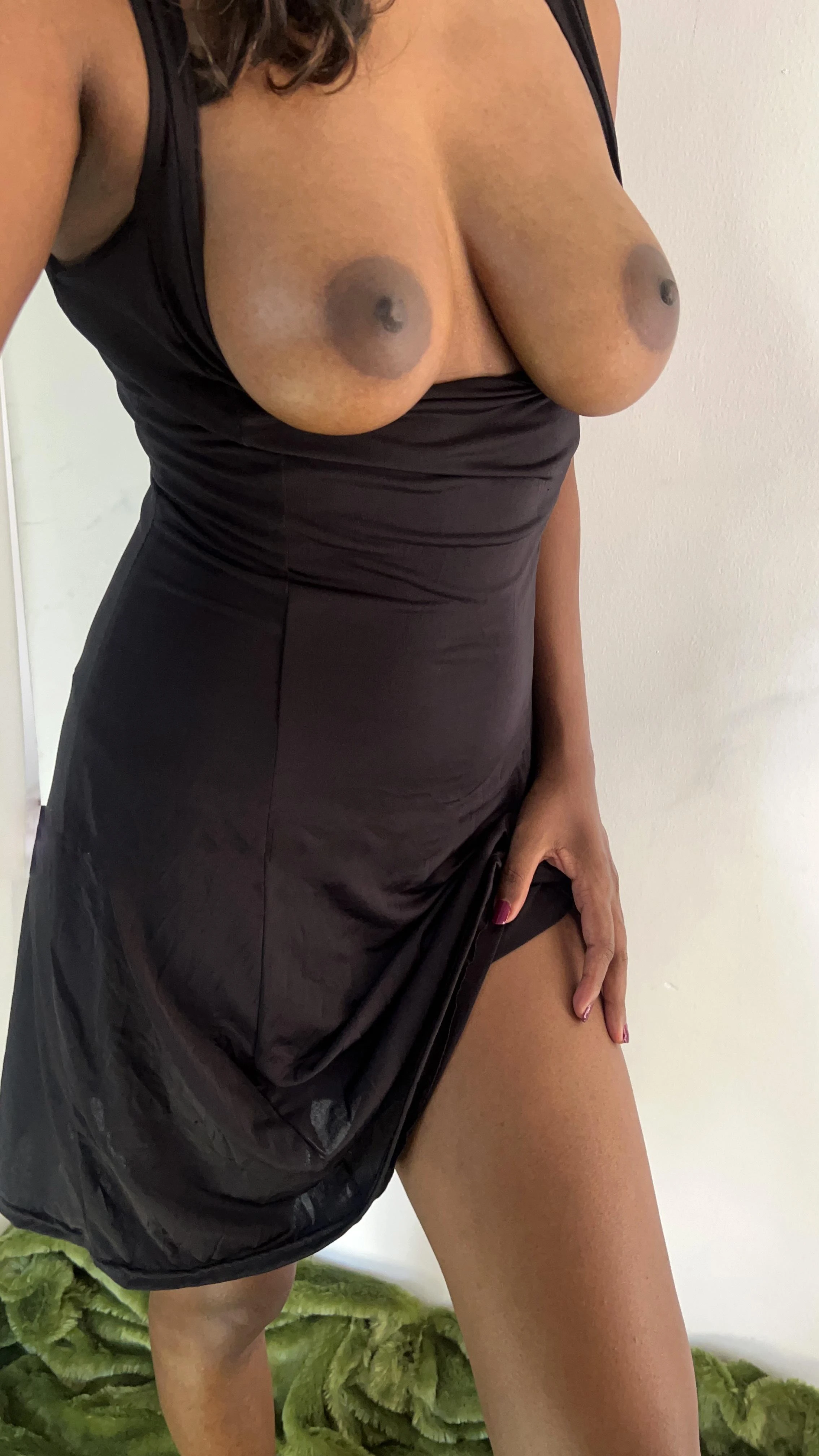 High-fashion study of Desi woman with large pendulous breasts showing black satin dress and green ruffled rug background with direct gaze