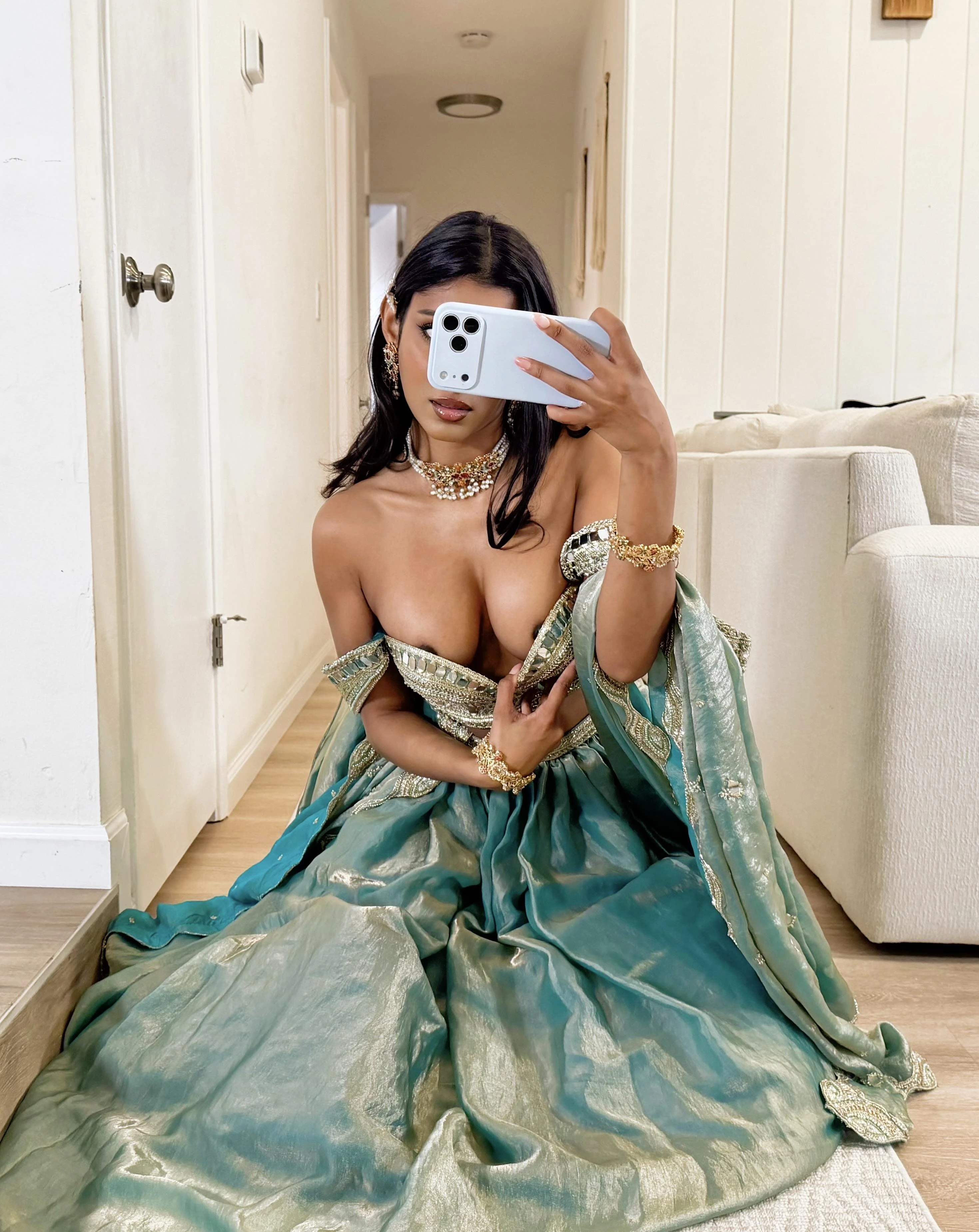 Desi lady with large exposed breasts in ornate teal-green gown kneels on light rug while holding smartphone to obscure face