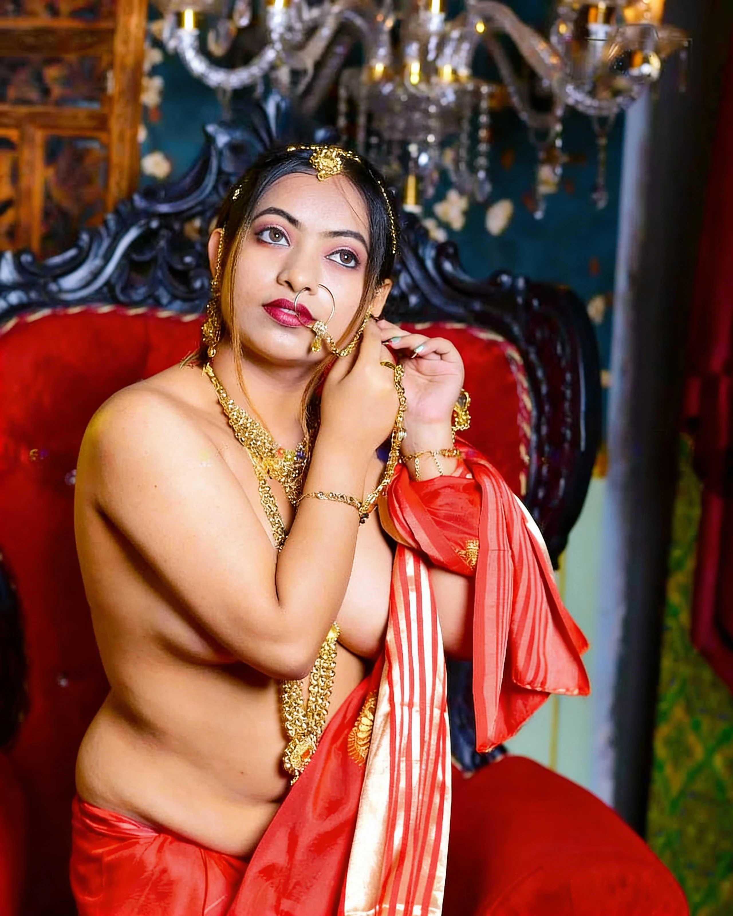 Desi woman with bare upper body wearing red saree and traditional gold jewelry seated on plush red chair in opulent setting