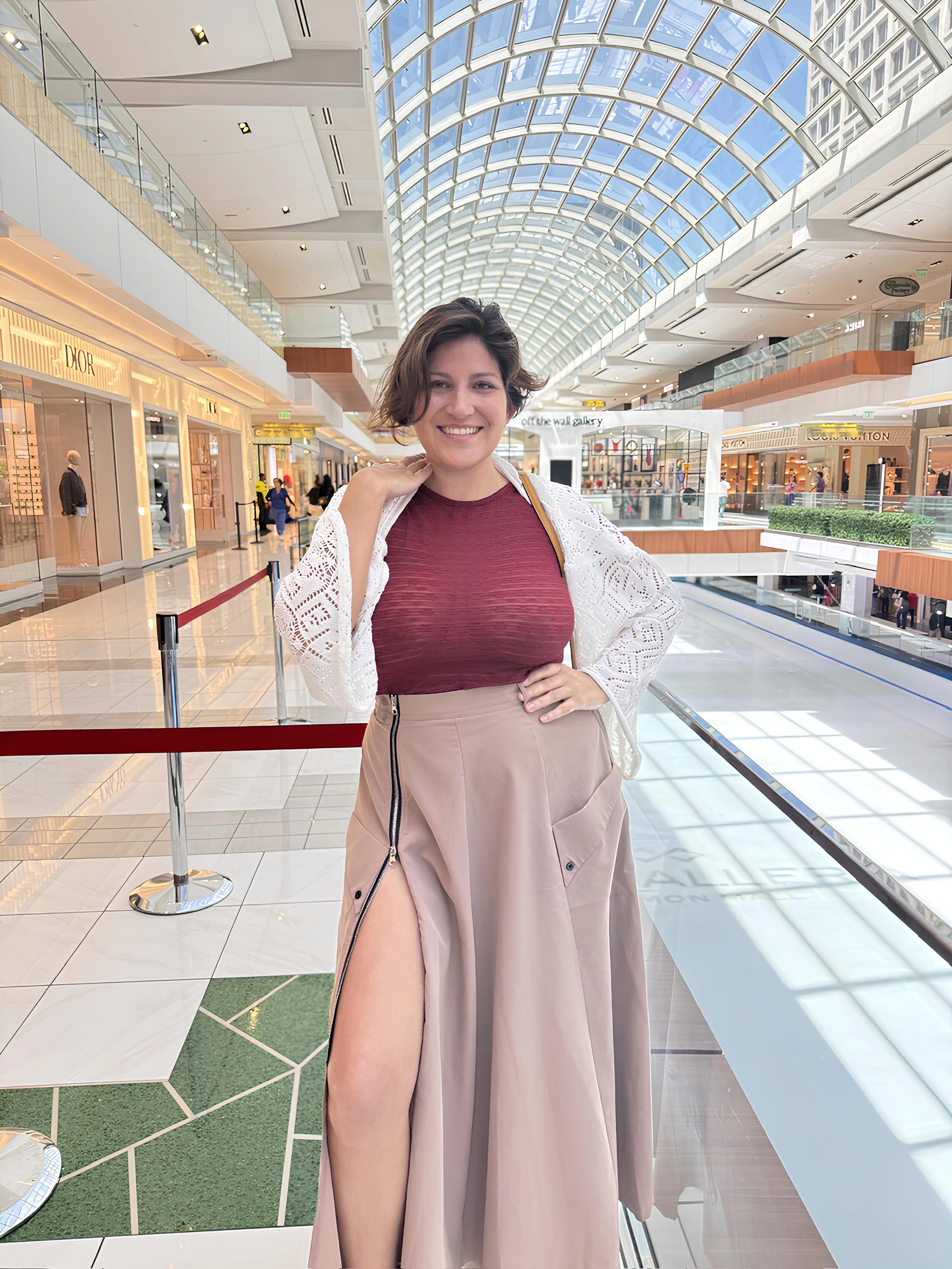 Woman with large full breasts in maroon form-fitting top and light brown high-waisted skirt with thigh-high slit posing in shopping mall with luxury storefronts visible