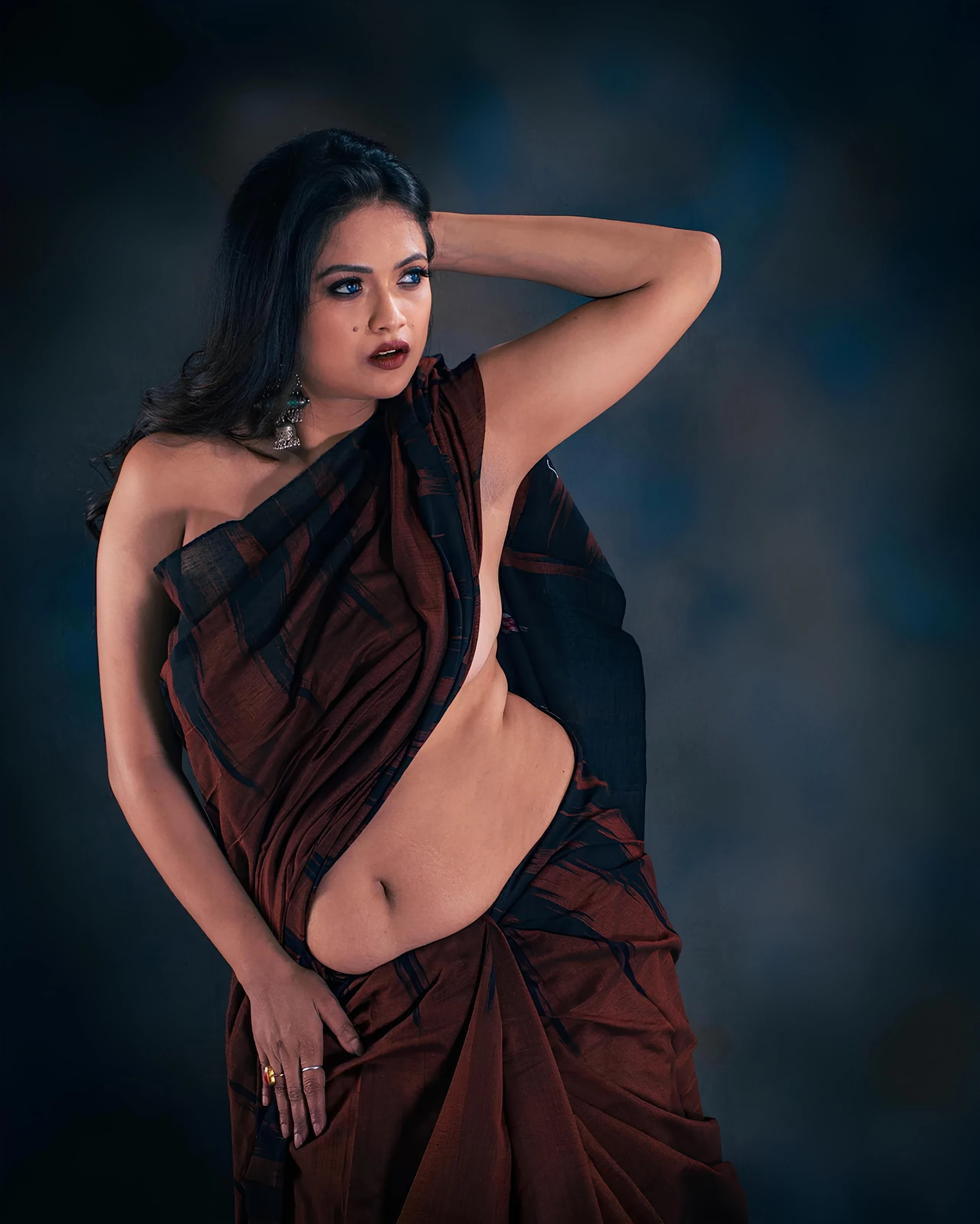Desi model in rust and black sari with striking blue eyes and burgundy lips against moody blue-gray gradient background with dramatic lighting