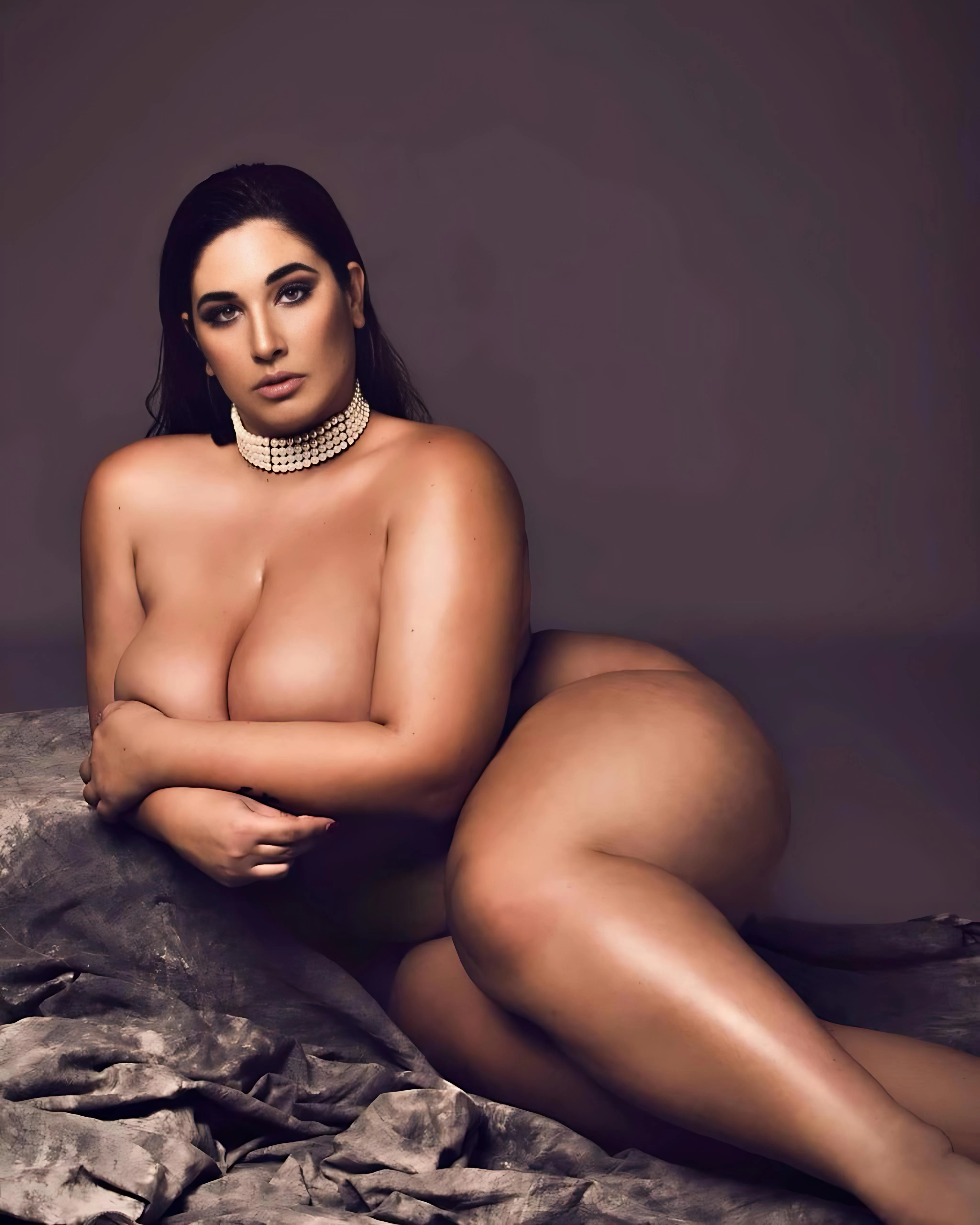 Full-body nude portrait of woman with full breasts wearing pearl choker necklace lying on dark gray fabric with dramatic lighting against muted gray background