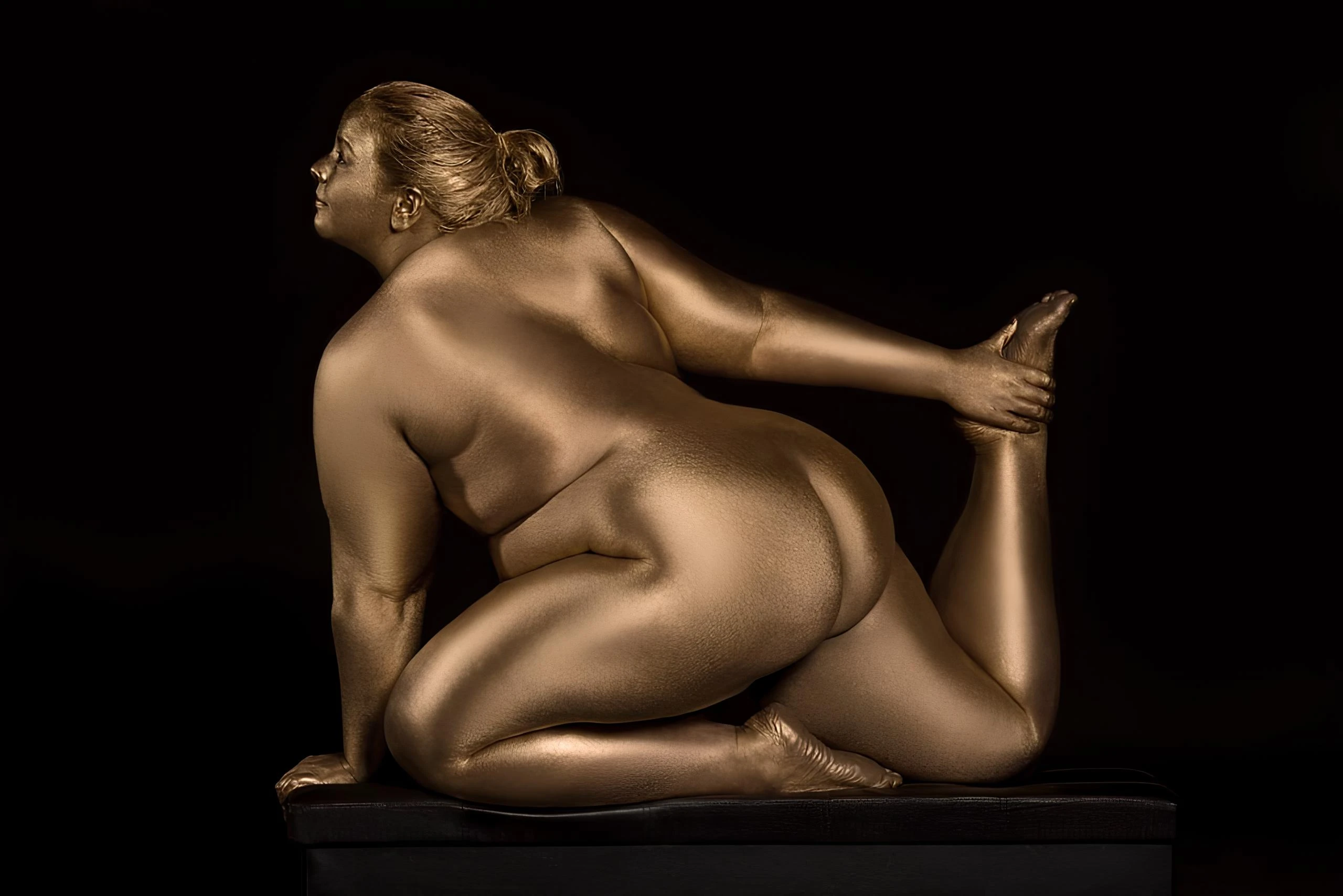 BBW model with large pendulous breasts and visible ass in golden body paint kneels on black surface